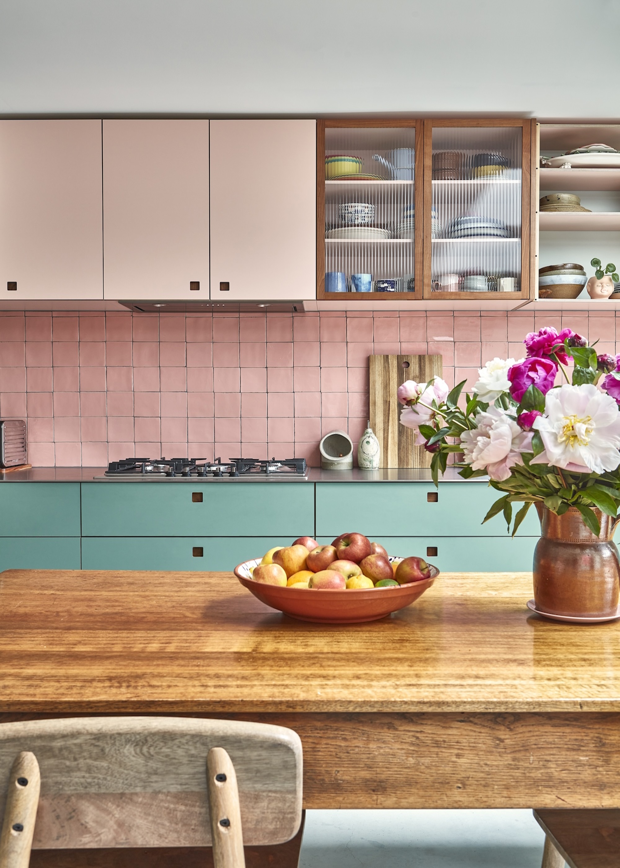 A coloful blue and pink kitchen with a dining table in the middle. On the dining table is a bowl of fruit as well as a large vase with flowers inside.