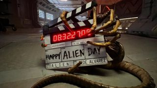 a spider-like alien holds a clapperboard on which is written "happy alien day"