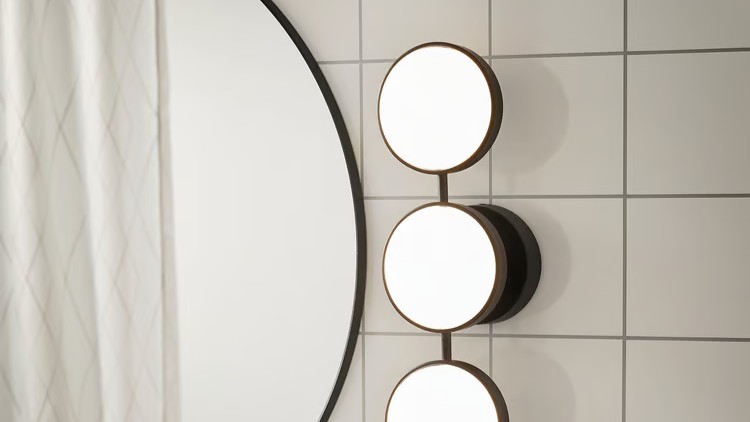 IKEA KABOMBA LED wall lamp in matt black with three circular frosted glass globes, mounted beside a round mirror in a white-tiled bathroom