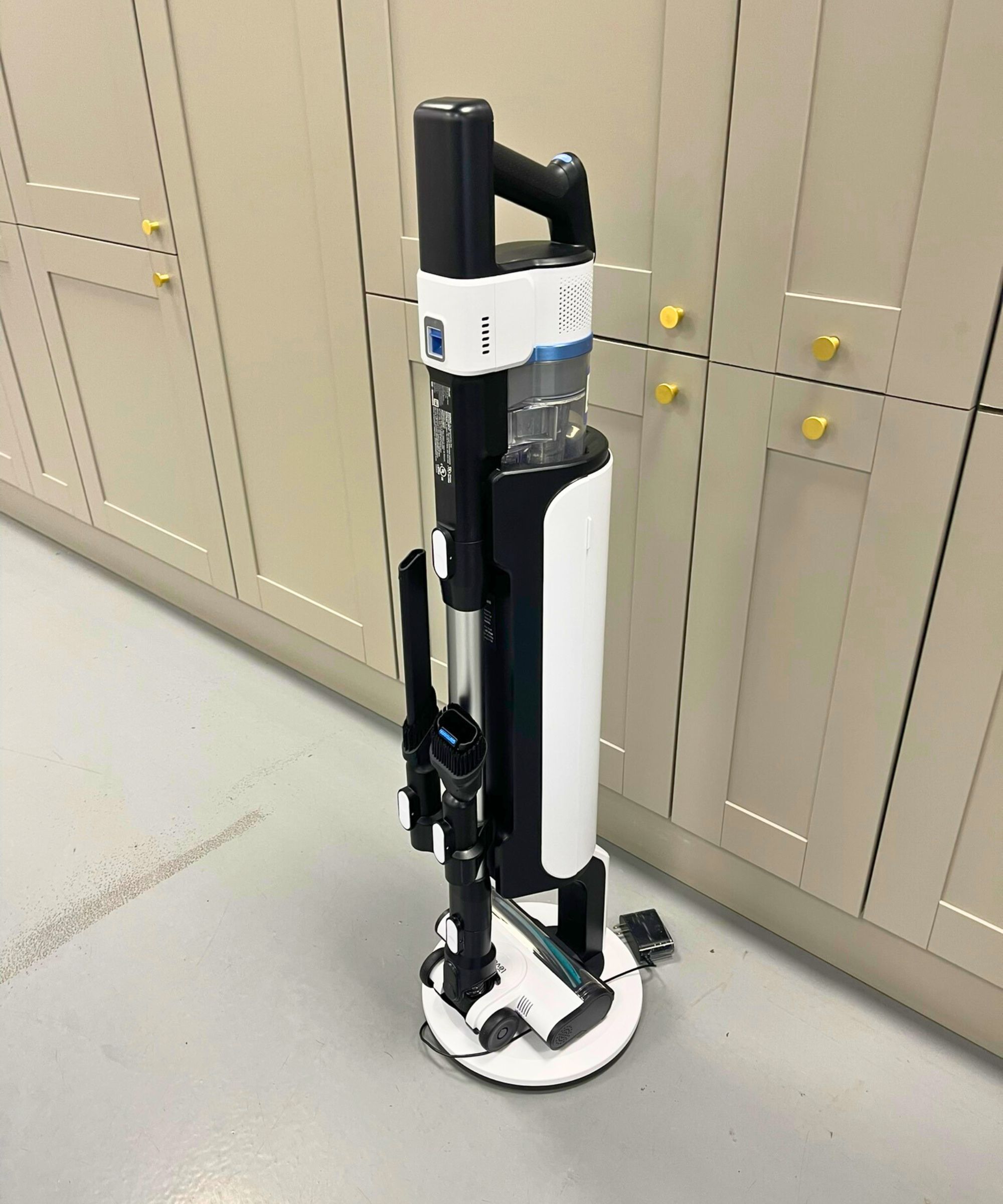Levoit AERO cordless vacuum cleaner docked on cleaning station, with two attachments attached to the vacuum's wand, on gray floor in front of gray cabinets with gold-colored handles.