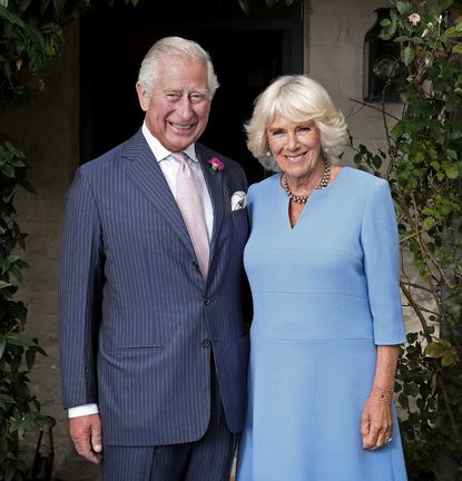 King Charles wearing a blue suit standing next to Queen Camilla, wearing a blue dress, in a garden outside 