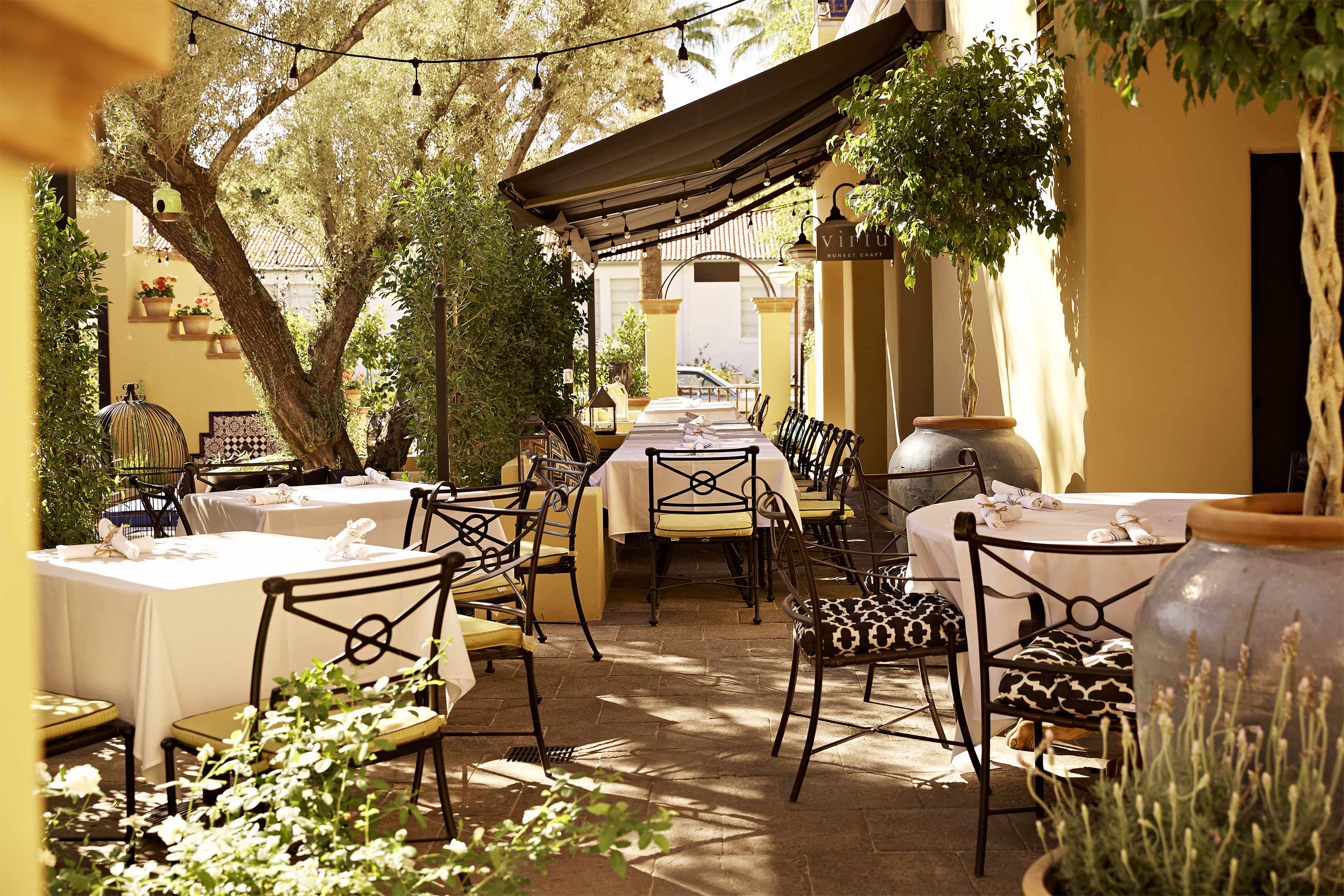 A sun-lit restaurant patio with wrought-iron chairs, white tablecloths, and yellow cushions is decorated with a number of thriving plants.
