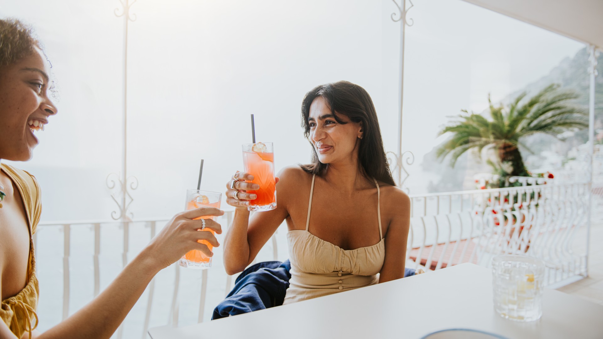 Two young women drinking pink-orange mocktails on a balcony toasting each other