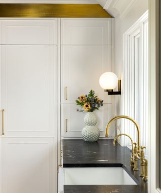 white kitchen with gold faucet, gold trim above cabinetry, granite countertops, butler sink, wall lights,