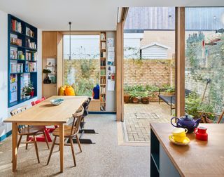 Kitchen-diner extension with wooden dining table and chairs, window seat, built-in blue-painted shelving and wooden corner sliding doors out to courtyard garden