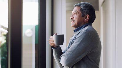 A man holds a coffee cup and looks thoughtful as he gazes out the window.