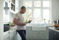 Man in his late 50s scrolling on his cell phone in a bright kitchen.