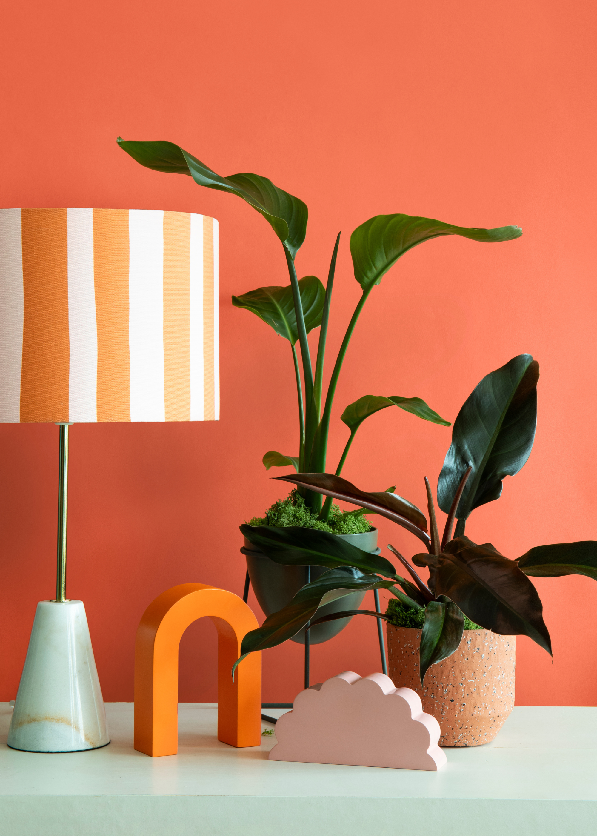 A striped lamp on a teal table by an arched orange decor piece and a pink cloud, beside a pair of potted plants against an orange wall