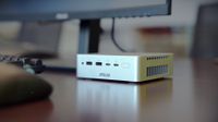 A compact ASUS Ascent QN10 mini PC sits on a desk beneath a monitor. The silver aluminium chassis features front‑facing ports including USB‑A, USB‑C, and a power button, highlighting its small 0.7‑liter design and workspace‑friendly footprint.