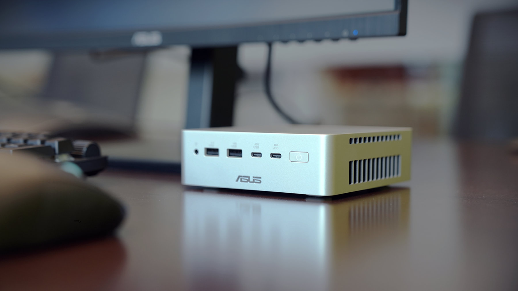 A compact ASUS Ascent QN10 mini PC sits on a desk beneath a monitor. The silver aluminium chassis features front‑facing ports including USB‑A, USB‑C, and a power button, highlighting its small 0.7‑liter design and workspace‑friendly footprint.