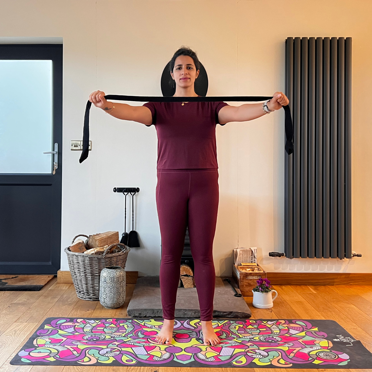Maysun Hassanaly performs a shoulder floss movement with a yoga strap. She is standing on a colorful yoga mat, holding the strap in front of her chest with wide arms.