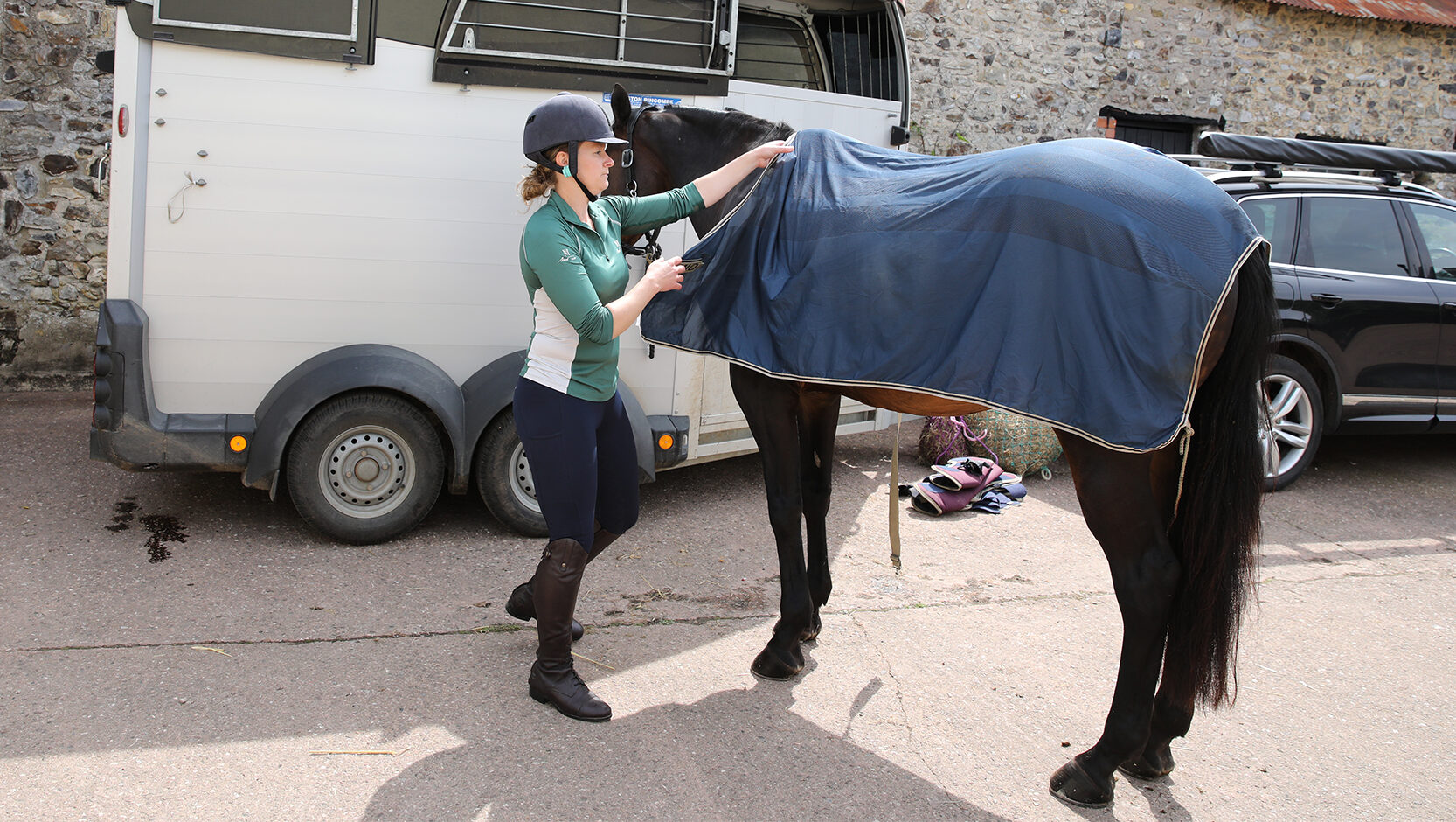 Horse standing outside a trailer with a lady taking off a cooler rug