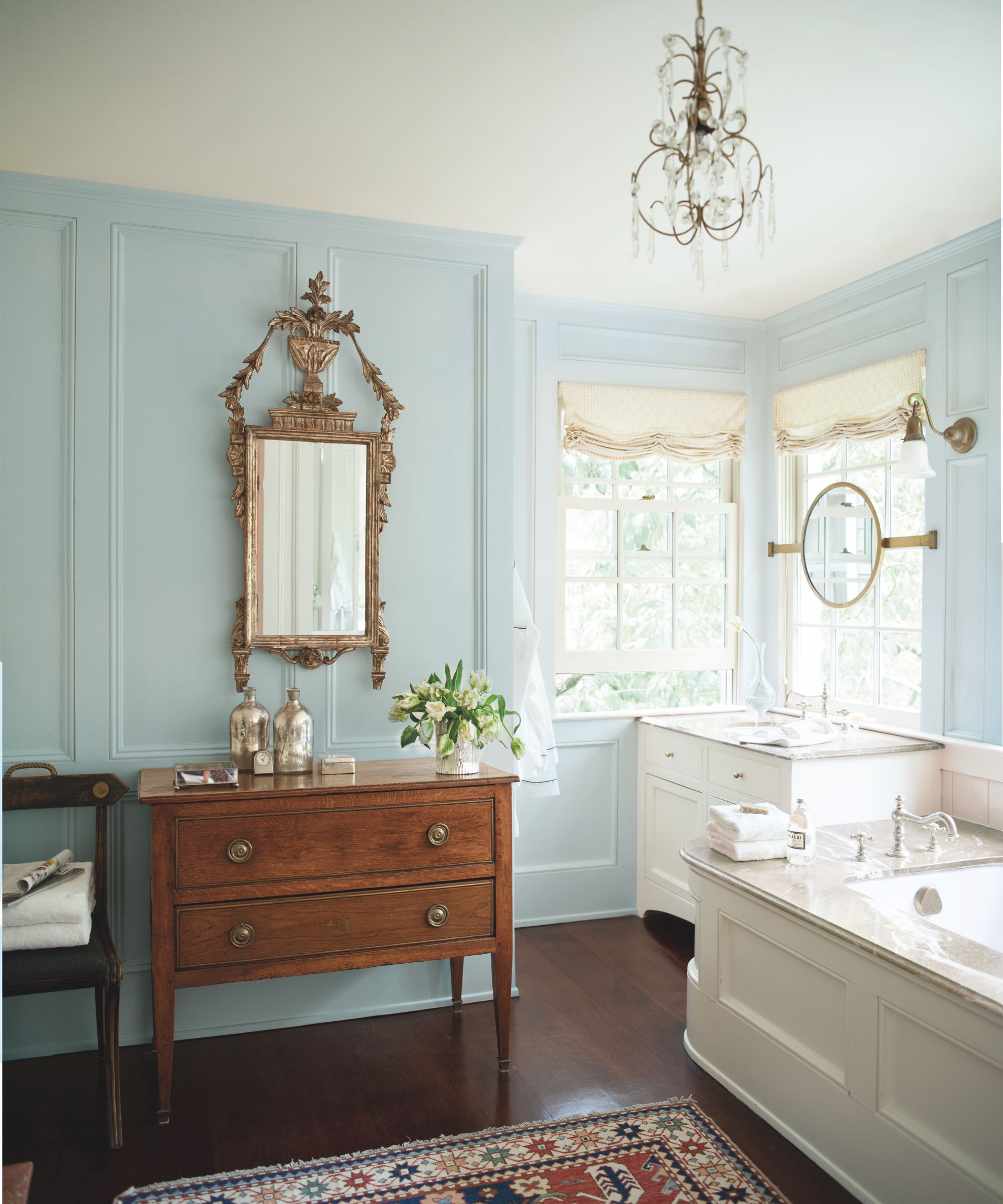 Traditional bathroom with pale blue painted walls, marble bathtub, windows with roman blinds, mahogany side table with decor and ornate gold mirror and Persian rug