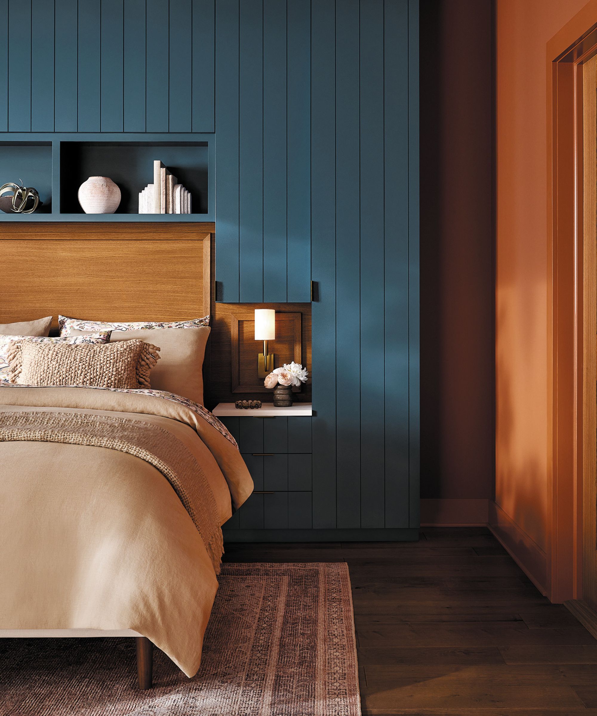 Bedroom with a teal panelled wall, a bed with a wooden headboard and neutral bedding, dark wood floors, an orange wall and doorway to the right, and alcoves holding books and trinkets.