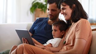 A mother, a father, and a child sit on the sofa as they look at a tablet
