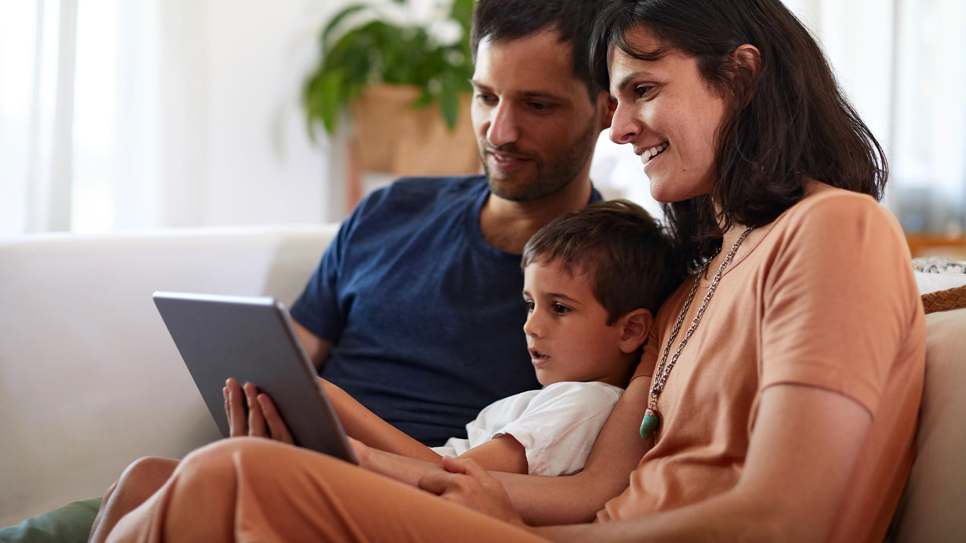 A mother, a father, and a child sit on the sofa as they look at a tablet