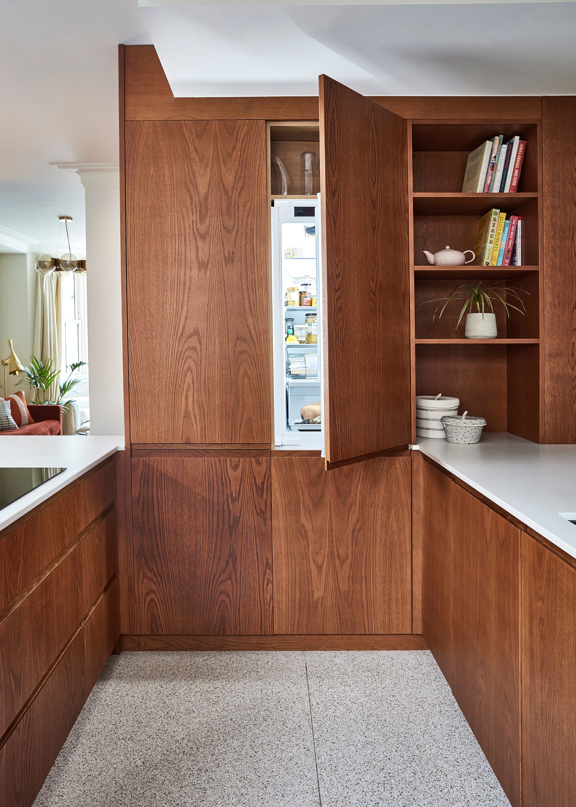 A kitchen with wooden features and cabinets. One section of the cabinets open to a fridge. There is also open shelving in another section with books and plants.