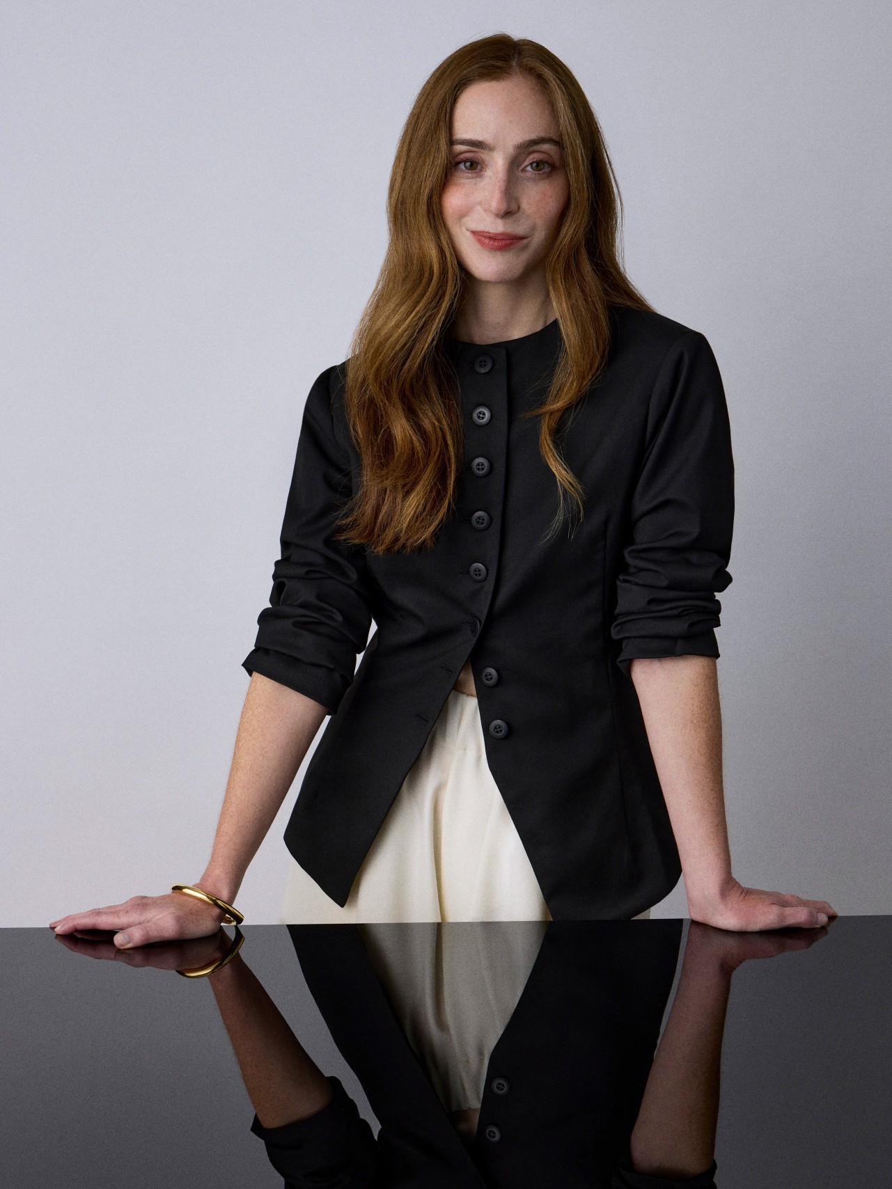 a woman with dark red hair wearing a black sweater and white pants behind a black desk