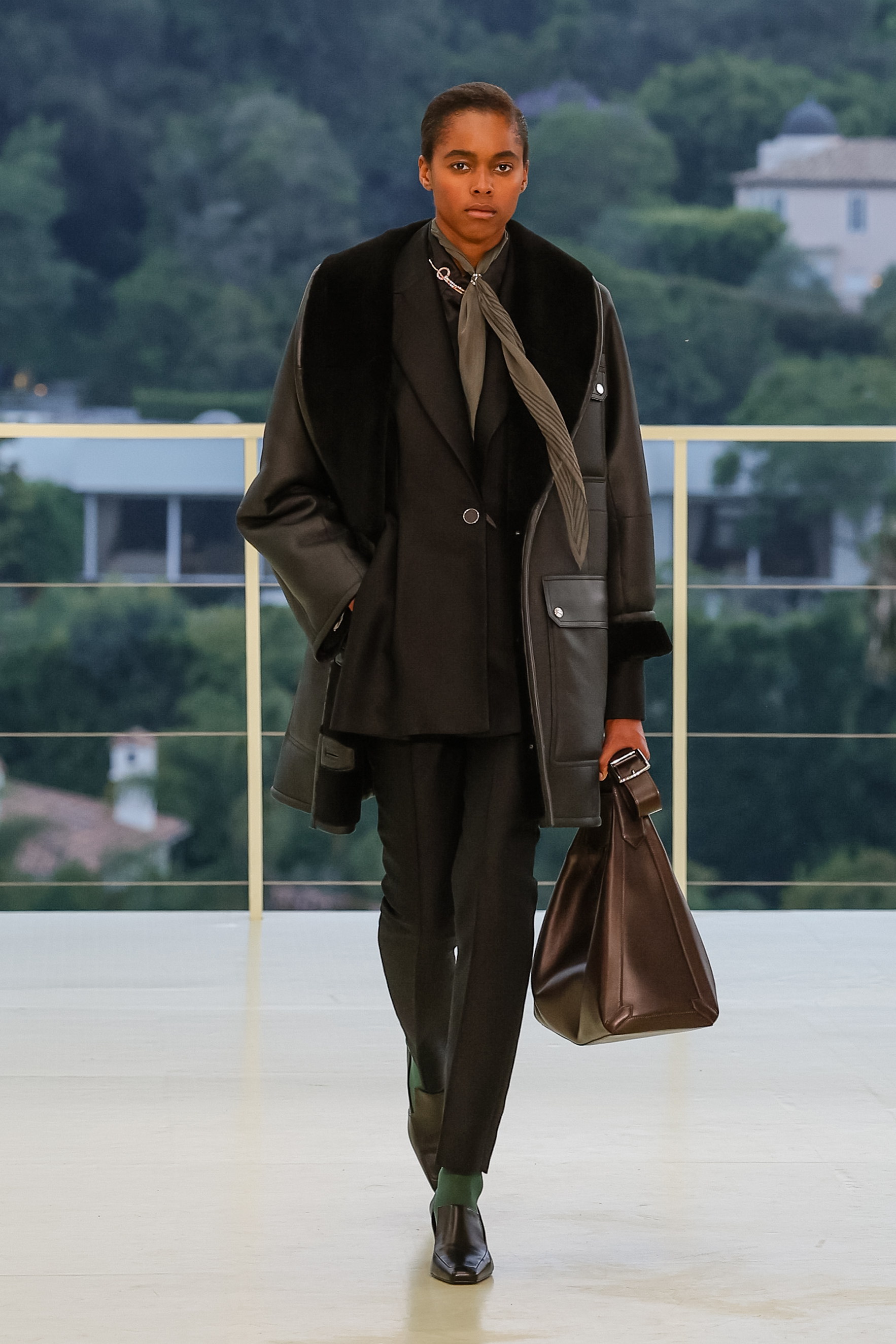 a model carries a brown bag and wears black pants with a black jacket.