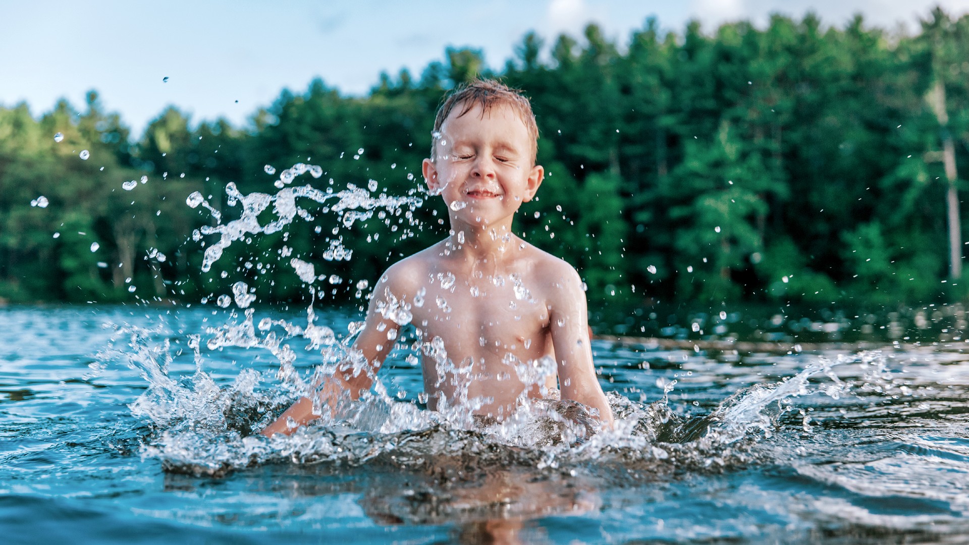 Young boy is shown splashing water in a lake. He has his eyes closed as he splashed. Woodland is shown in the background behind the lake