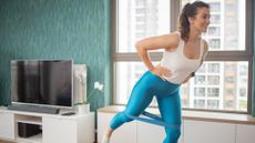 Woman exercising in an apartment setting. She has a resistance band wrapped around her knees and lifts one leg behind her. She wears a tank top and blue three-quarter length leggings. 