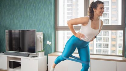 Woman exercising in an apartment setting. She has a resistance band wrapped around her knees and lifts one leg behind her. She wears a tank top and blue three-quarter length leggings. 