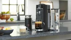 Nespresso VertuoPlus on a granite countertop with the aeroccino, a cappuccino and fruit bowl beside it