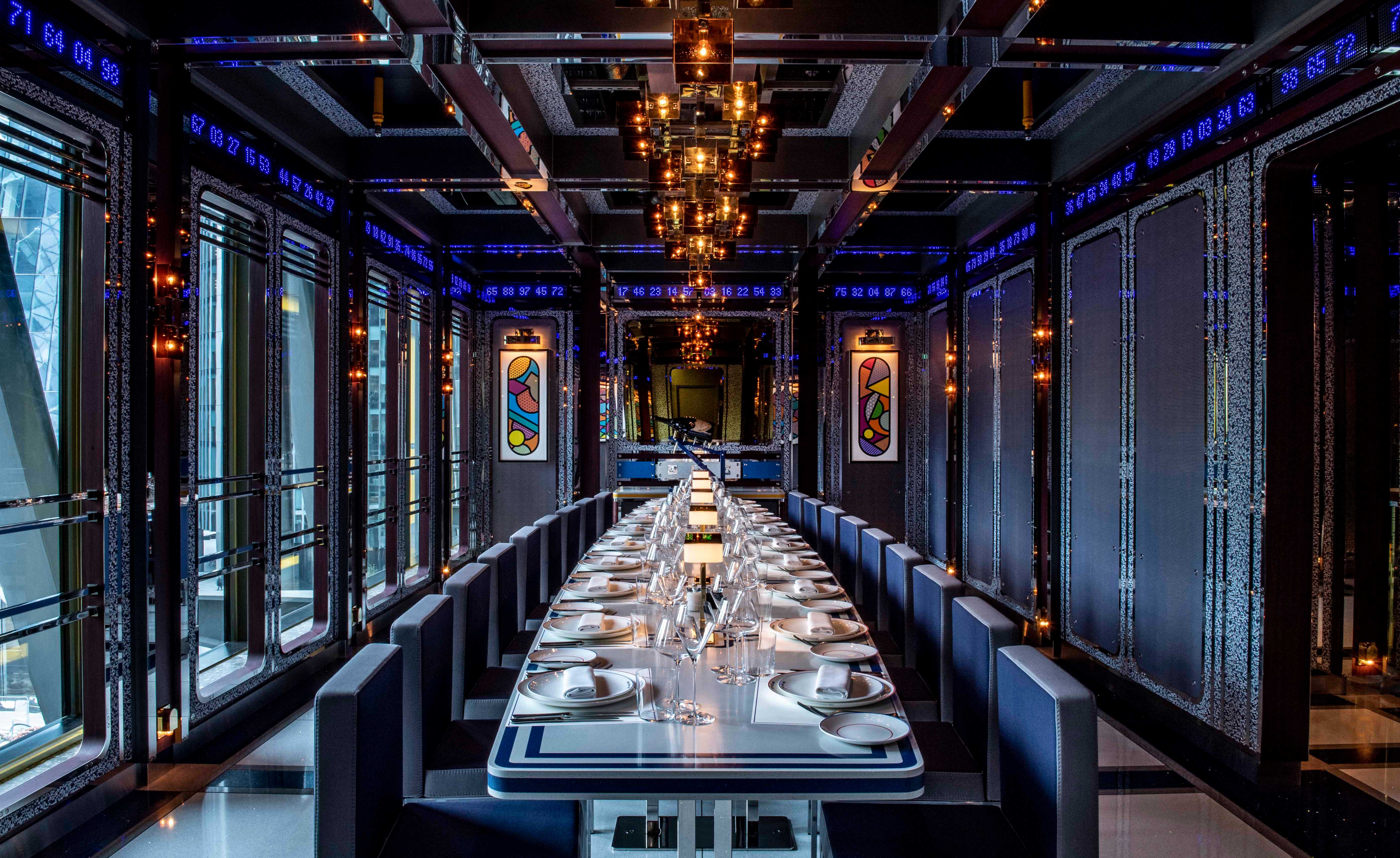 Dining room at Bob Bob Cité restaurant with a long white and blue table, blue covered chairs, a chandelier, large windows and a strip of glowing blue numbers running below the ceiling.