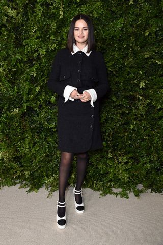 Selena Gomez stands in front of a foliage wall wearing a black button up dress with black tights