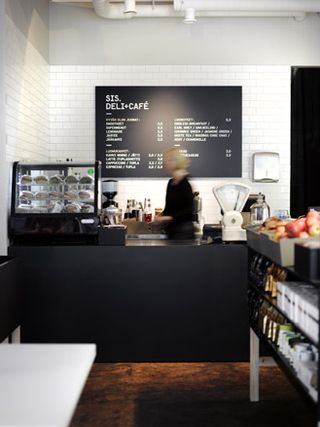 SIS Deli & Cafe, Finland. A cafe with a black serving counter with a glass food display case on it, shelves with products on them and a menu on the wall.