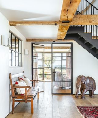 Large hallway with wooden beams, white painted walls, wooden flooring, rug, bench, elephant statue, black metal and glass doors leading on to living room, black metal staircase,