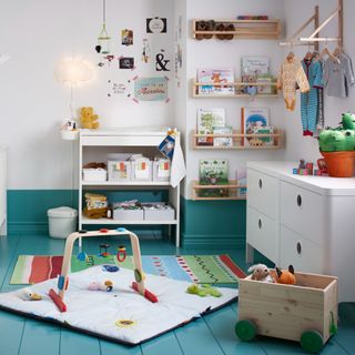 White changing table with nappies and other items stored underneath, white chest of drawers on blue floor with rug and toys