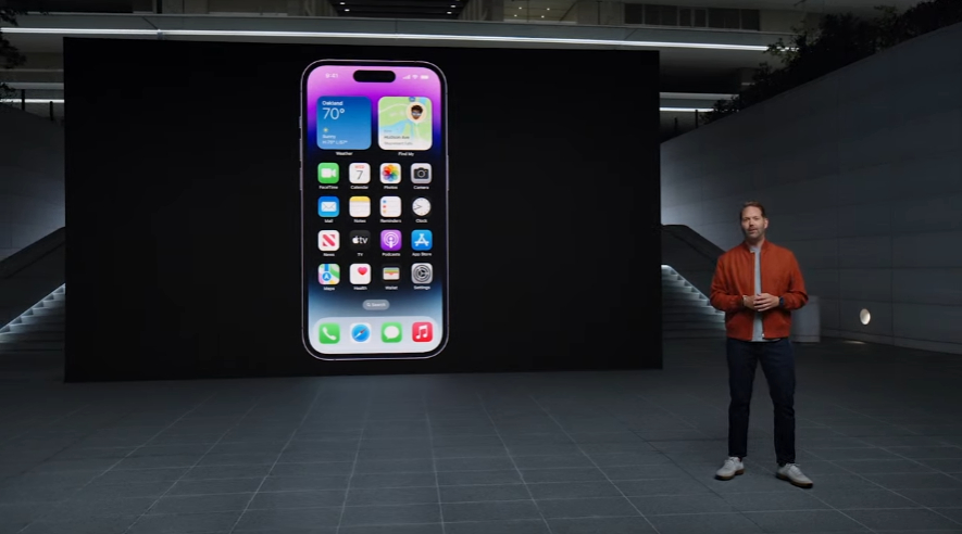 A person ina red jacket stands in front of a giant screen showing the iPhone 14