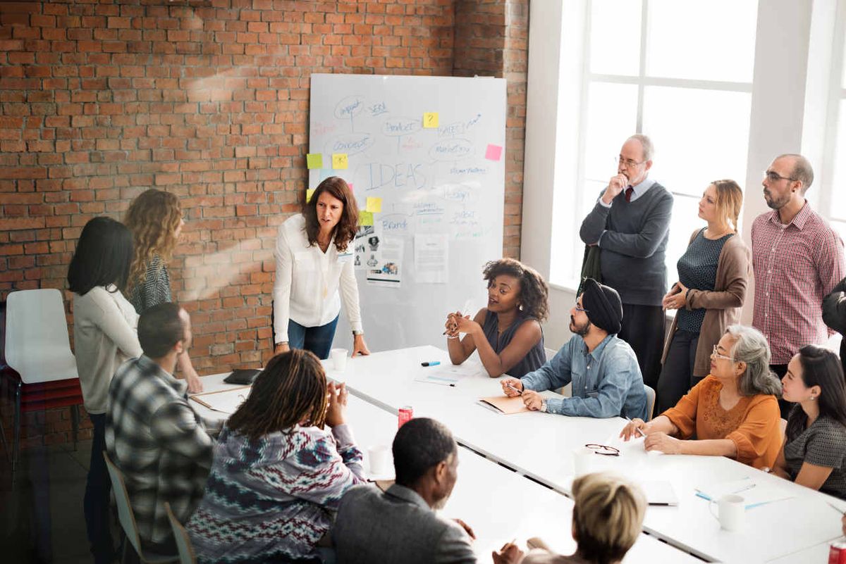 A group of diverse men and women sit at a conference table or stand while discussing meeting topics.