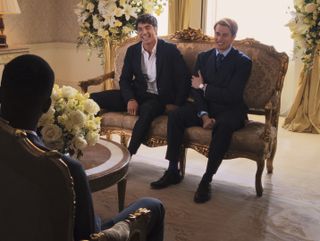 Alex (Taylor Zakhar Perez) and Henry (Nicholas Galitzine) are sitting on an ornate, classical two-seater chair and giving false smiles for a television interview. Henry is rubbing his upper right arm.