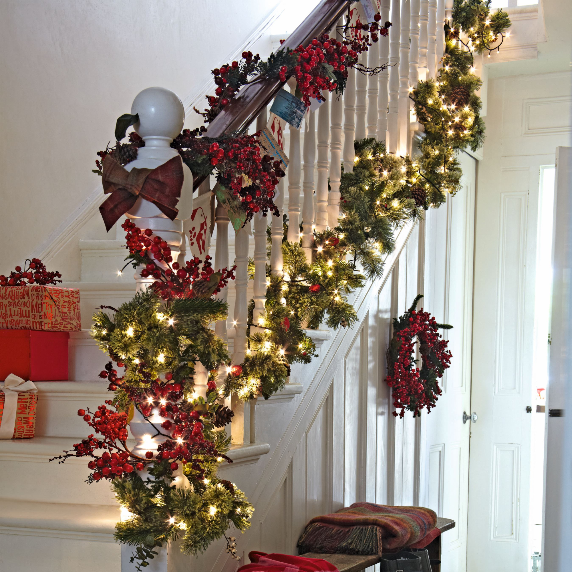 Festive hallway and staircase, period style staircase with bannisters, real fir tree foliage garland with red berries and fairy lights, art deco tiled floor, wooden bech with scarves and wellies, berry wreath, cupboard under stairs