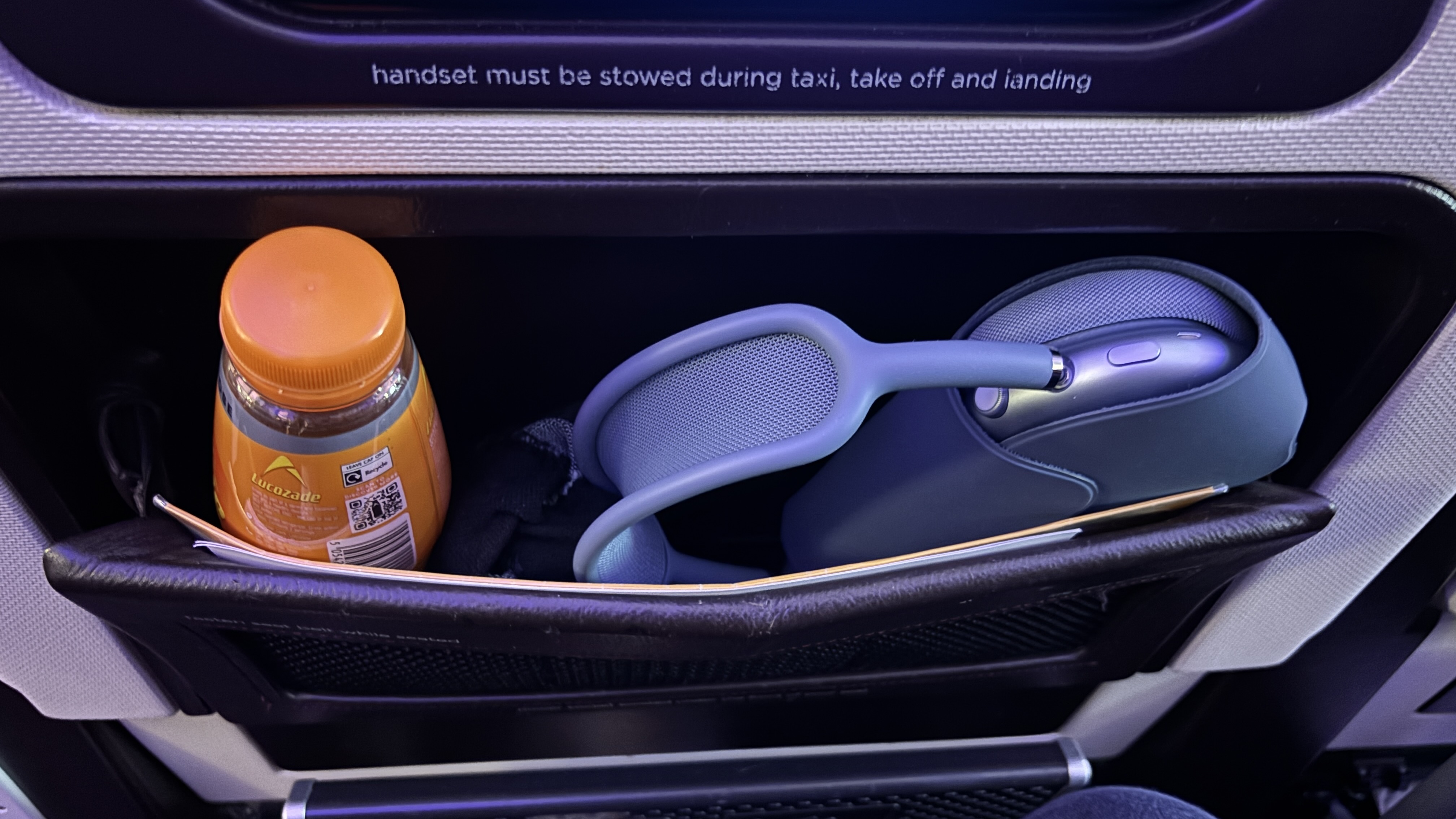 AirPods Max sowed away in the seat pocket on an aeroplane with a bottle of Lucozade next to them