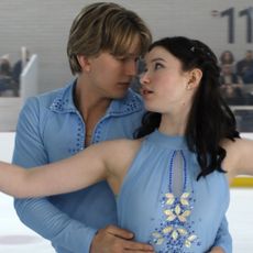 Cale Ambrozic as Brayden Elliot and Madelyn Keys as Adriana Russo wearing matching blue uniforms figure skating together with a crowd in the bleachers in a still from finding her edge