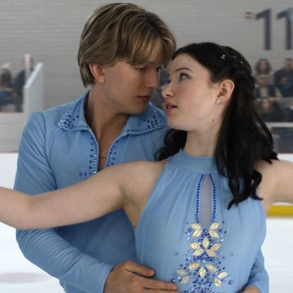 Cale Ambrozic as Brayden Elliot and Madelyn Keys as Adriana Russo wearing matching blue uniforms figure skating together with a crowd in the bleachers in a still from finding her edge