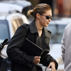 Gigi Hadid steps out in New York, showcasing a dramatic grey wool coat paired with a beige turtleneck and white sneakers. Carrying a black bag and laptop, the model completes the chic look with sleek hair and dark sunglasses.