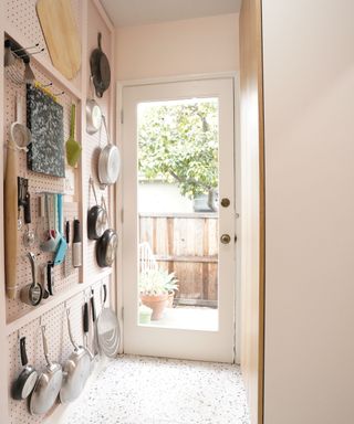 A pegboard kitchen storage setup on a light pink wall in a hallway