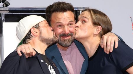 Getty image of Ben Affleck in a suit being kissed on the cheek by Kevin Smith and Jason Mewes in 2019. Smith has on signature backwards baseball cap.