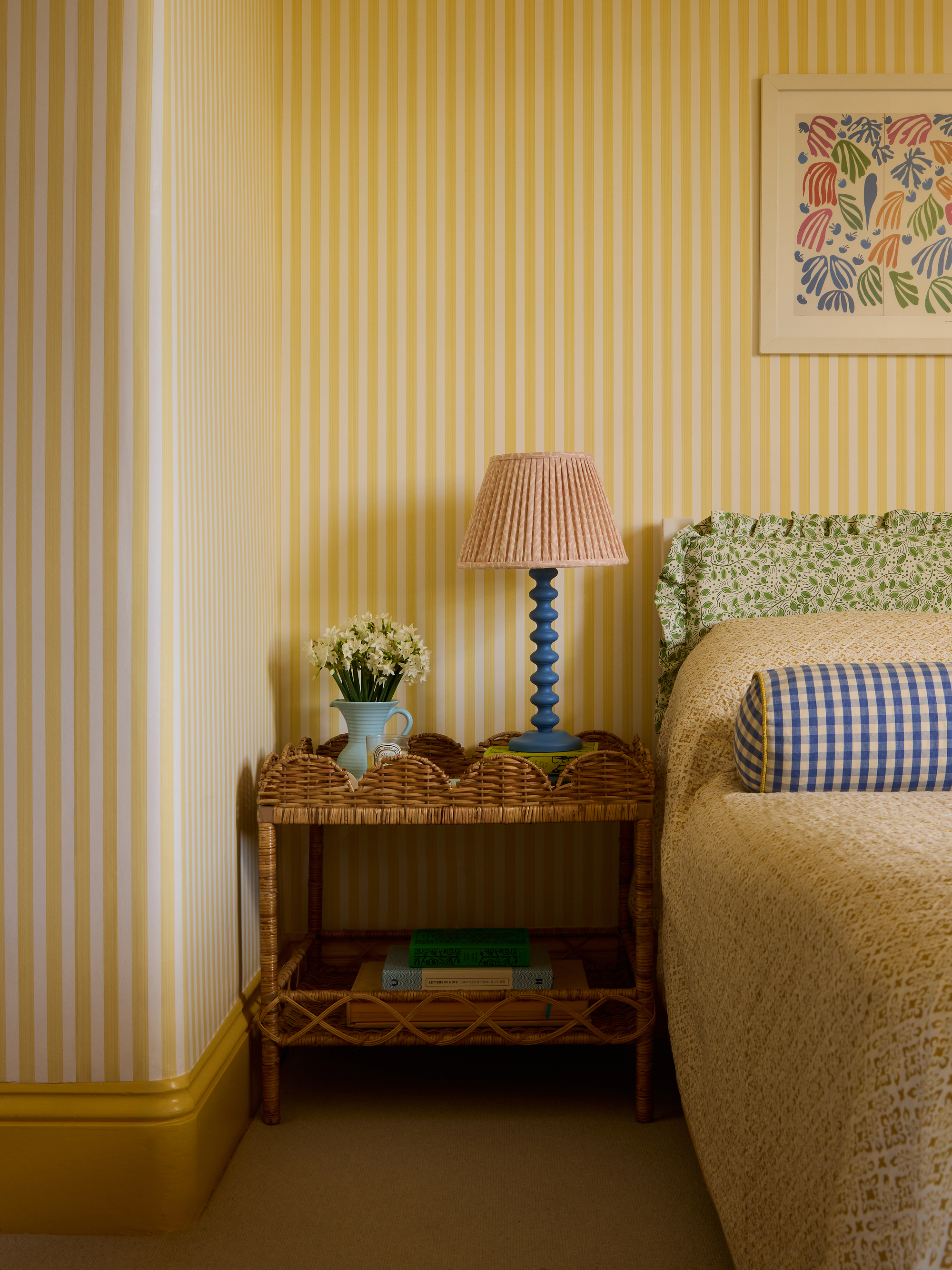 yellow bedroom with striped wallpaper, yellow bedding with gingham bolster and frilly floral headboard, bobbin lamp with rattan side table with scalloped edge and vase of flowers