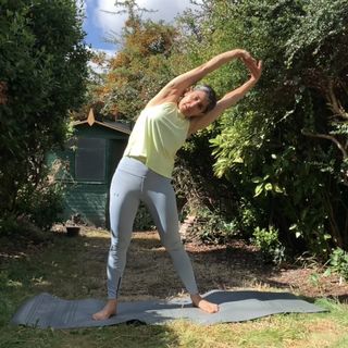 woman stretching sideways in yoga pose yellow top light blue leggings garden background