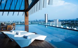 View of the rooftop at AC Hotel, Guadalajara, Mexico featuring wooden decking, a pool, white geometric style lounge chairs and blue and wooden chairs. There are also views of surrounding buildings