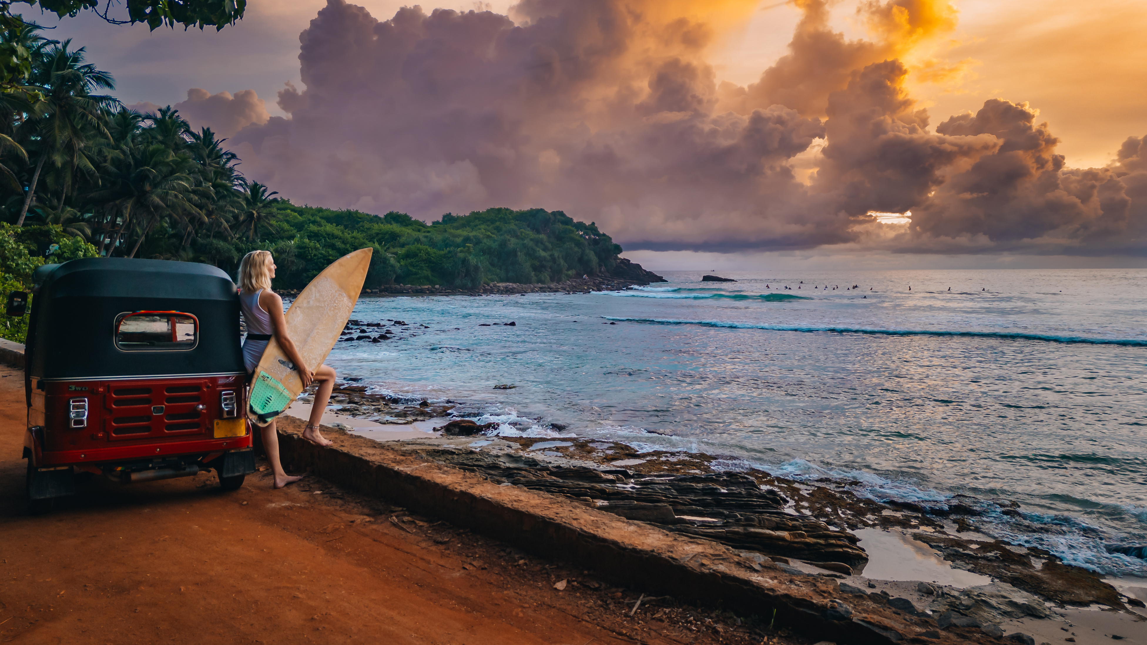 A blonde woman holding a surfboard leans against a tuk-tuk in front of the water at Hiriketiya Beach