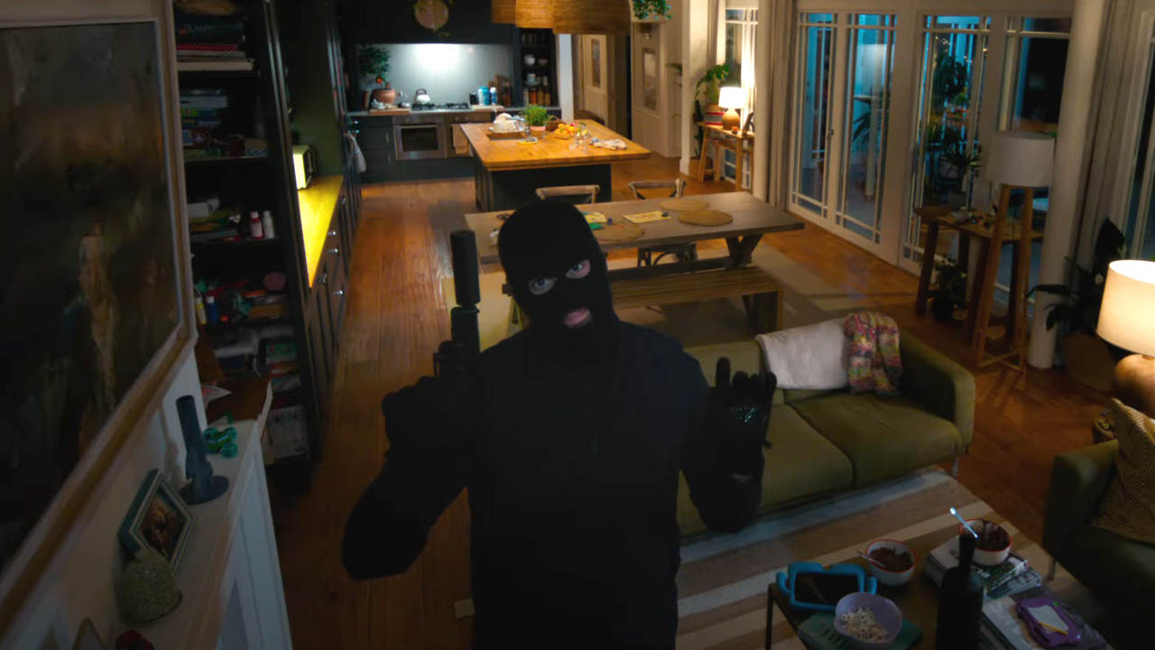 A masked attacker waves to a home security camera while standing in the living room of Drop.