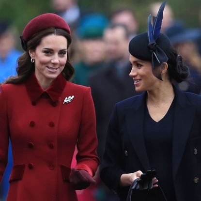 Kate Middleton wears a red coat with a fur collar and a matching hat, Meghan Markle wears a navy coat and matching dress, Prince Harry wears a white shirt, blue tie, and large overcoat