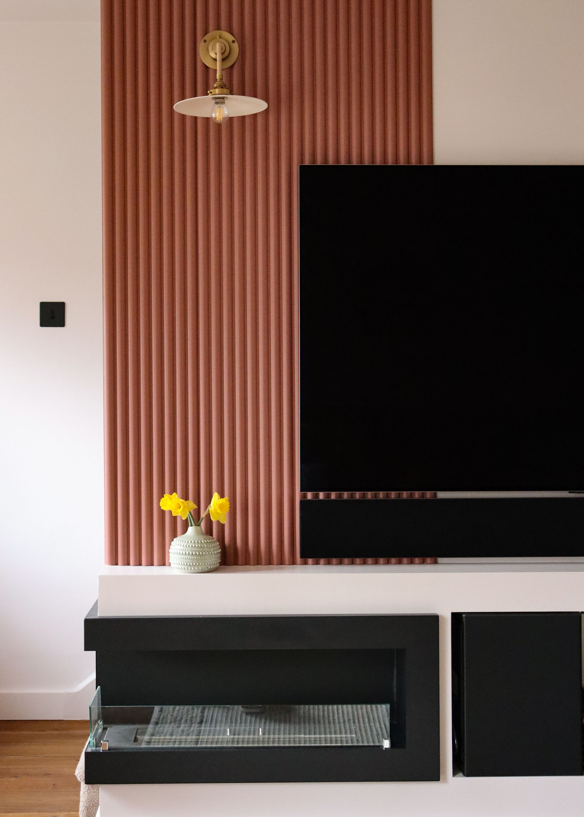 A tv with with extra storage at the bottom. There is also wall panels places to create depth and dimension. On the shelf below the tv is a plant in a vase