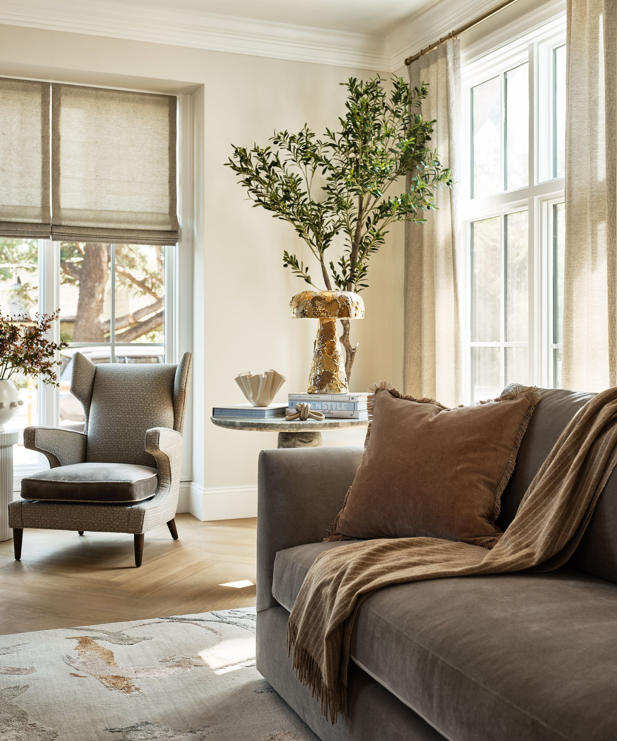 An elegant, neutral-toned living room featuring a grey velvet sofa with a tan throw, a classic wingback armchair by the window, and a small marble side table with a gold mushroom lamp and a potted olive tree.